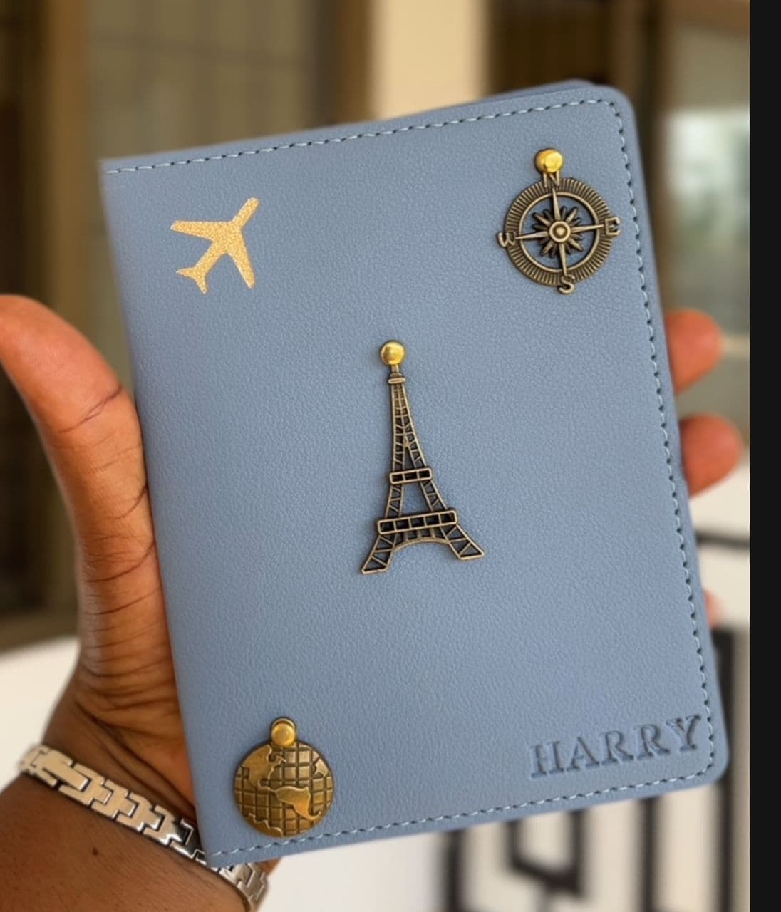Personalised light-blue P.U leather passport cover with a gold airplane, compass, Eiffel Tower and globe charms, embossed HARRY, held in hand