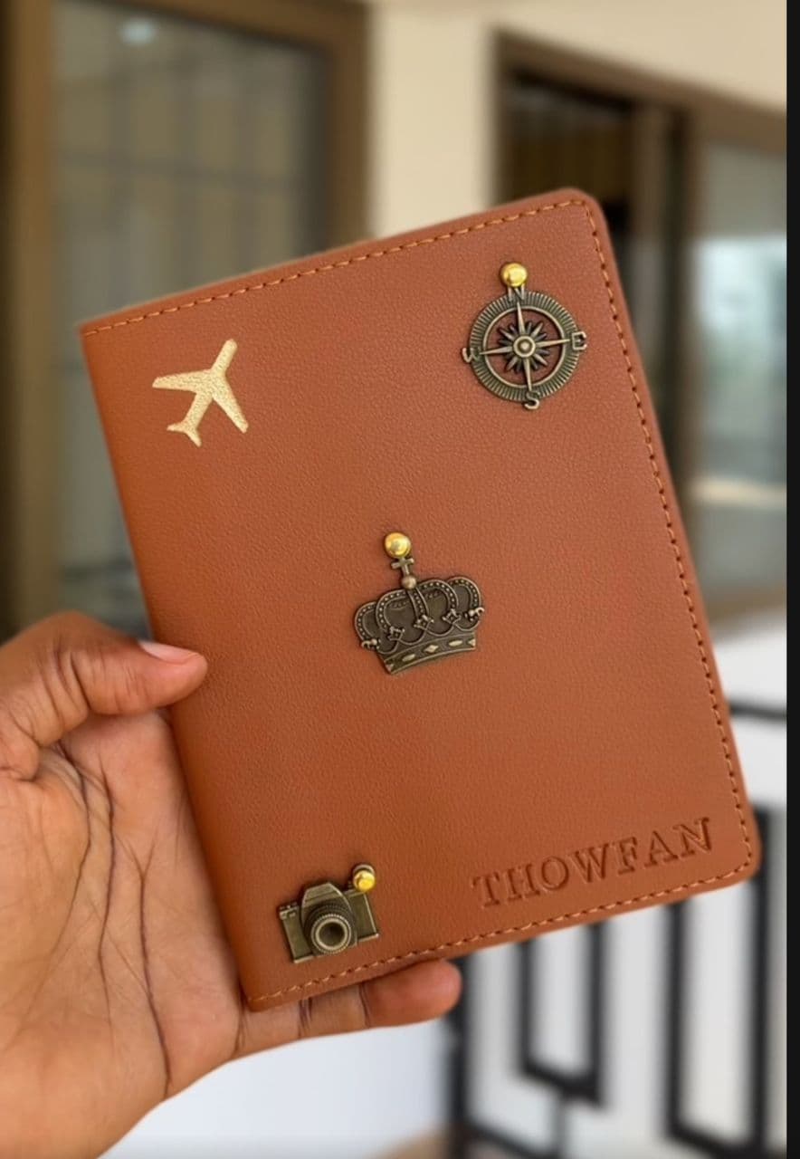 Personalised tan passport cover with airplane, compass, crown and camera gold charms, embossed THOWFAN, held in hand